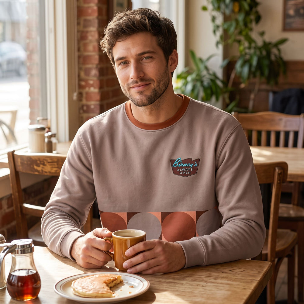 Relaxed adult model wearing Birney's Always Open Sweatshirt in cozy living room setting with warm natural lighting.