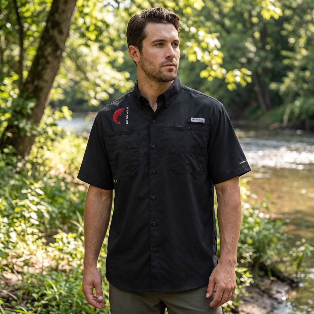 Male model wearing black Columbia PFG Chinook Guide Shirt with embroidered Chinook graphic, chest pockets, and rod loop on a riverbank scene