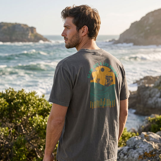 Casual male model wearing a distressed vintage maritime graphic t-shirt in an outdoor natural setting with greenery and water.