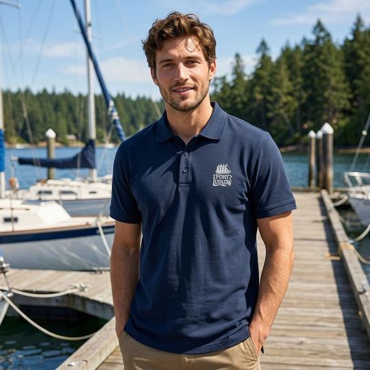 Adult male model in navy polo with sailing vessel embroidery, outdoors with lush greenery and open sky.