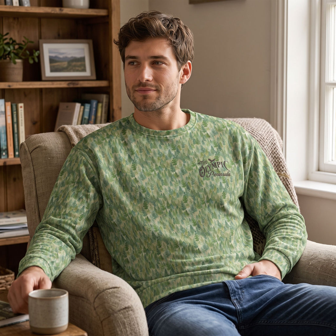 Relaxed man in Olympic Pines sweatshirt sitting in cozy living room near window with warm natural light.