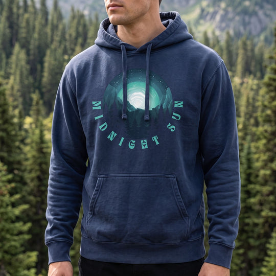 Close-up of Midnight Sun hoodie highlighting the celestial sky and mountain silhouettes graphic against a blurred outdoor background with greenery and sunlight.