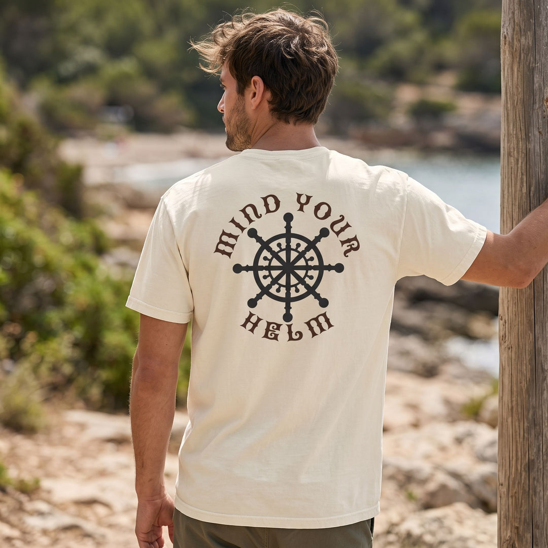 Casual male model wearing a navy garment-dyed T-shirt with ship steering wheel graphic outdoors amidst greenery and water.