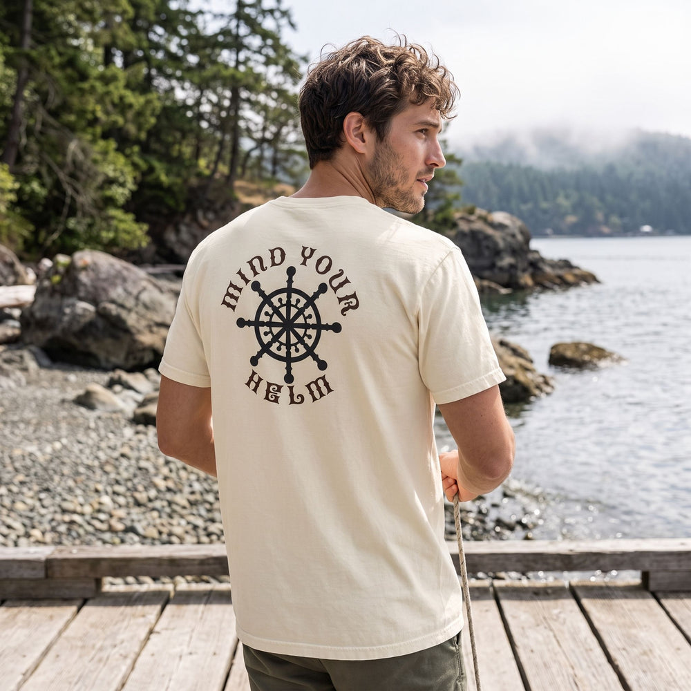 Relaxed fit men's T-shirt with maritime steering wheel graphic worn outdoors by a model with natural styling and background of blurred foliage.