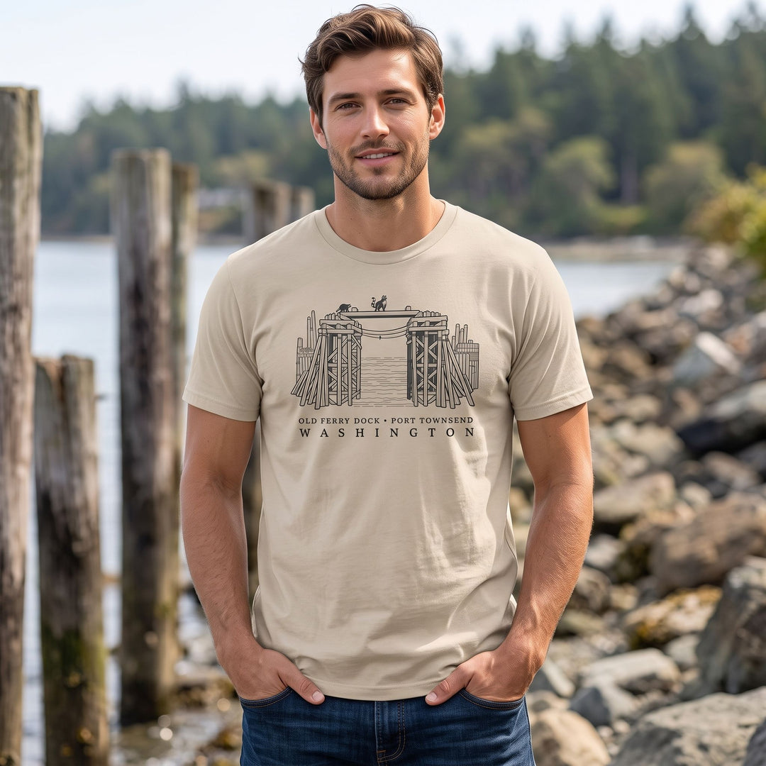 Model sporting the organic tee featuring coastal line art with raccoons, set against a natural outdoor background with foliage and water.