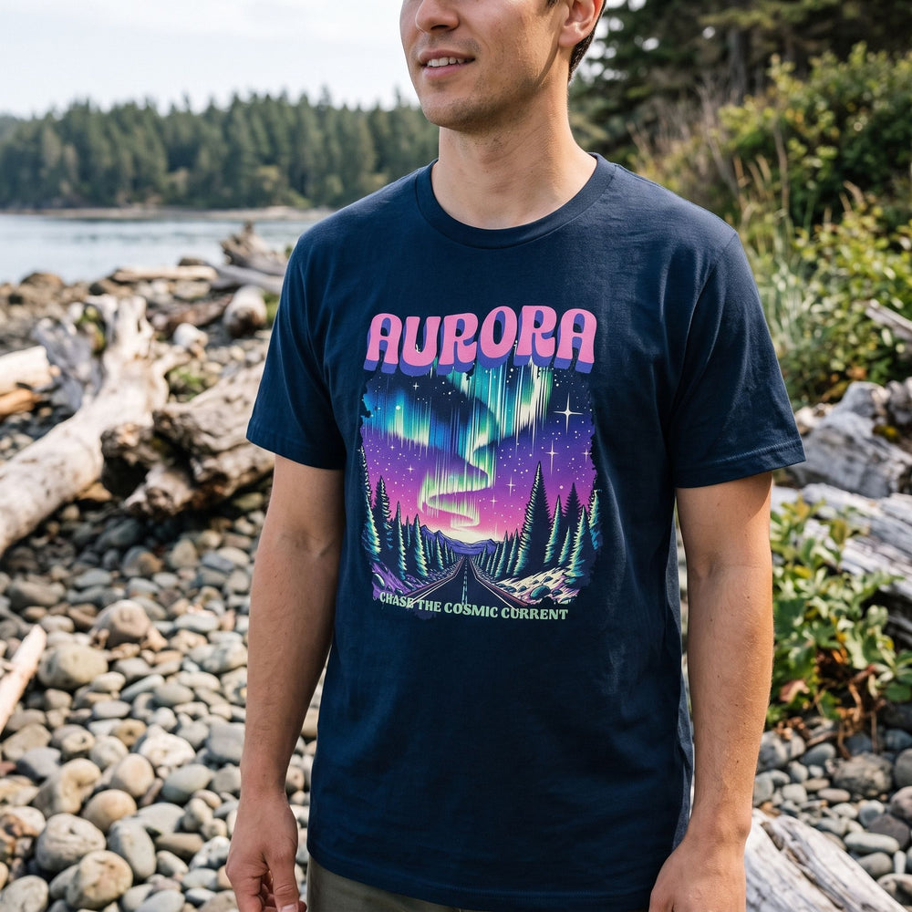 Person wearing Aurora Chase Tee in an outdoor setting near a rocky shoreline with greenery and water, showcasing the cosmic Aurora Borealis print.