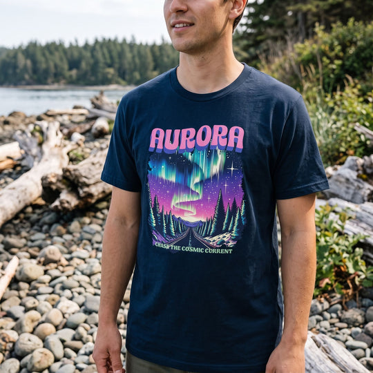 Person wearing Aurora Chase Tee in an outdoor setting near a rocky shoreline with greenery and water, showcasing the cosmic Aurora Borealis print.