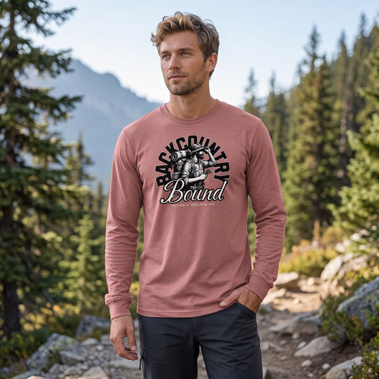 Adult male model wearing The Backcountry Bound Long Sleeve Tee in outdoor setting with greenery, rocks, blurred foliage, emphasizing rugged, nature-inspired style.
