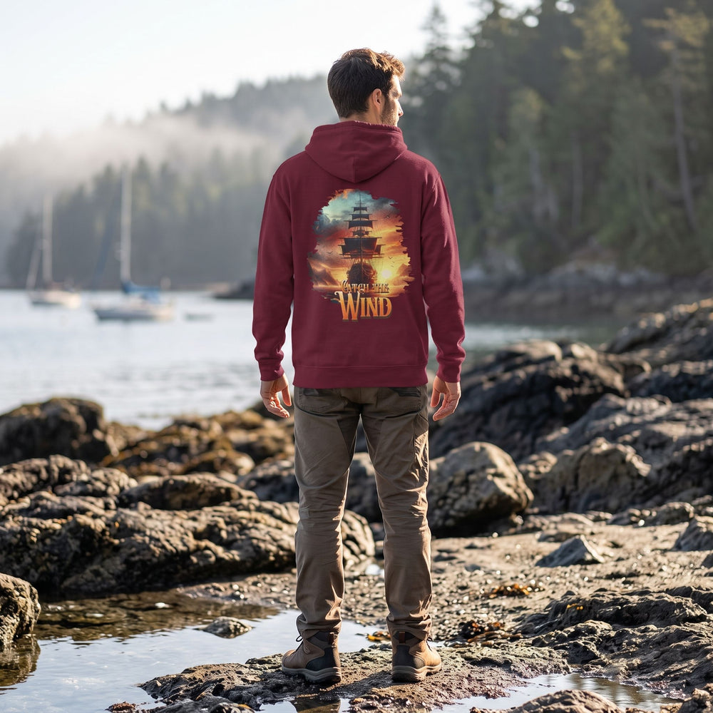 Casual male model wearing 'Catch the Wind' hoodie outdoors on rocky shoreline with greenery, emphasizing the rugged, adventurous style of the streetwear.