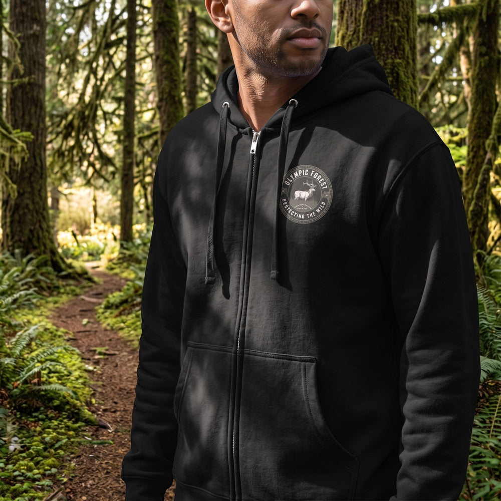 Model wearing The 1897 Olympic Forest Premium Zip Hoodie on a mossy trail among tall trees in a natural forest scene with dappled sunlight and blurred foliage in the background.