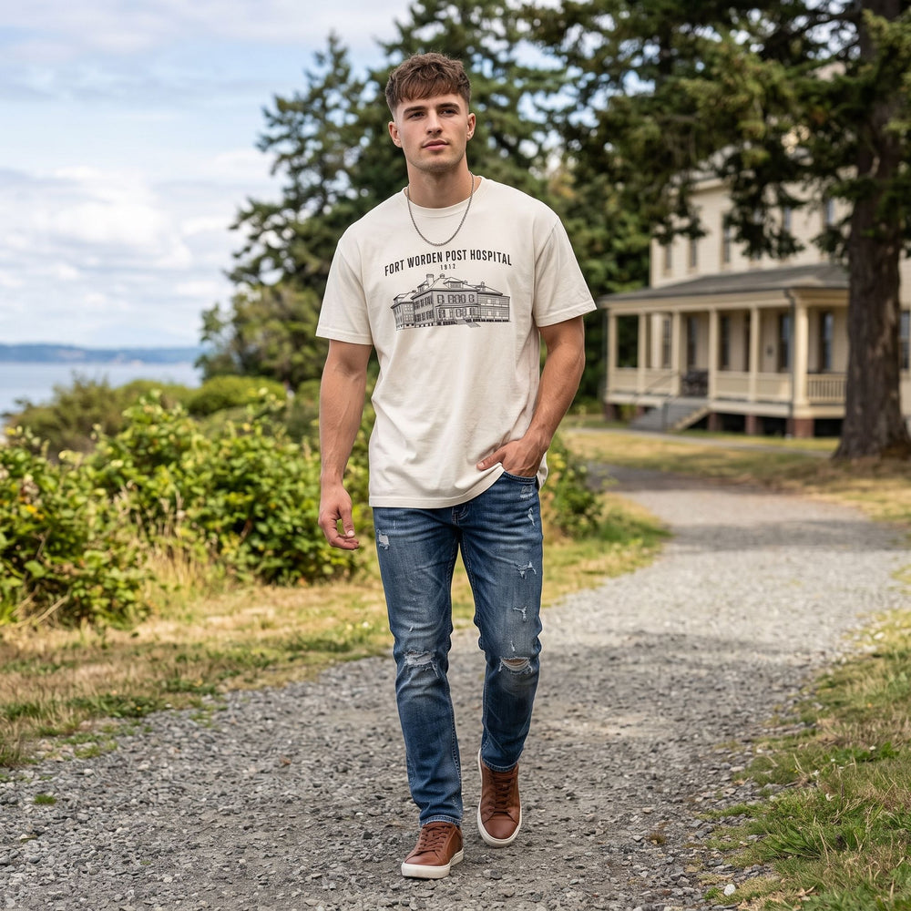 Young man modeling oversized heavyweight t-shirt with Fort Worden hospital graphic, outdoor scene with blurred natural background and sunlight.