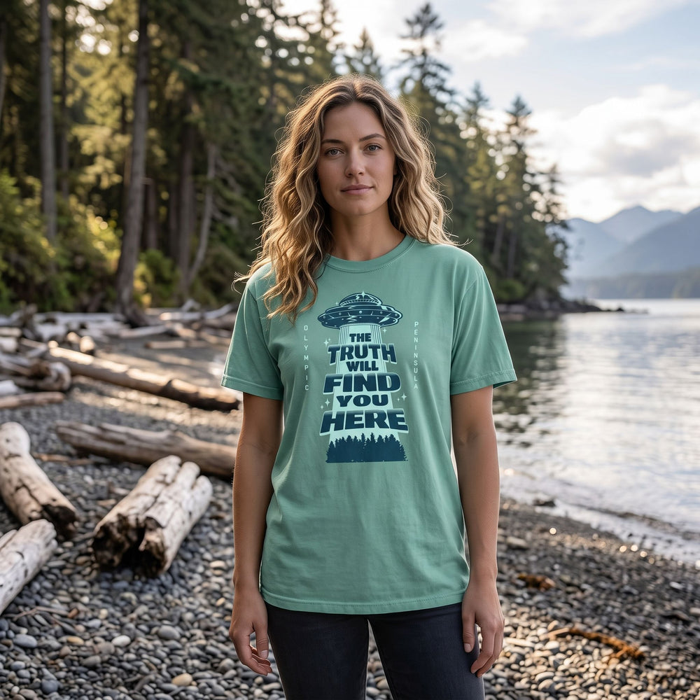 Casually dressed model wearing 'The Truth Will Find You Here' UFO tee outdoors on a forest trail, surrounded by greenery and natural sunlight.