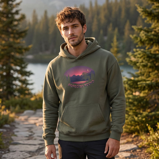 Model wearing Paradise Found Organic Mid-Weight Hoodie in outdoor setting with greenery, natural daylight, relaxed pose, and blurred foliage background.