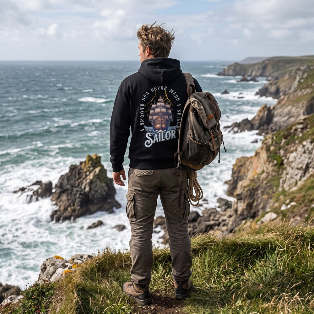 Rugged male model wearing The Skilled Sailor Heavyweight Hoodie on a windy coastal cliff with rocky coastline and churning sea in the background.