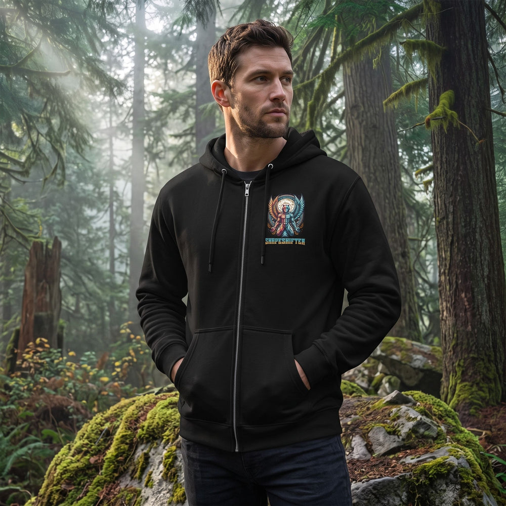 Male model wearing the Shapeshifter Premium Full Zip Hoodie with cosmic winged graphic, standing on rocky outcrop in misty forest at dawn showing outdoor adventure style.
