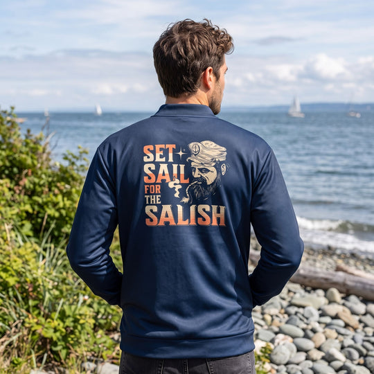 Adult model wearing Salish Sea inspired bomber jacket in outdoor setting with blurred natural foliage, rocks, and water.