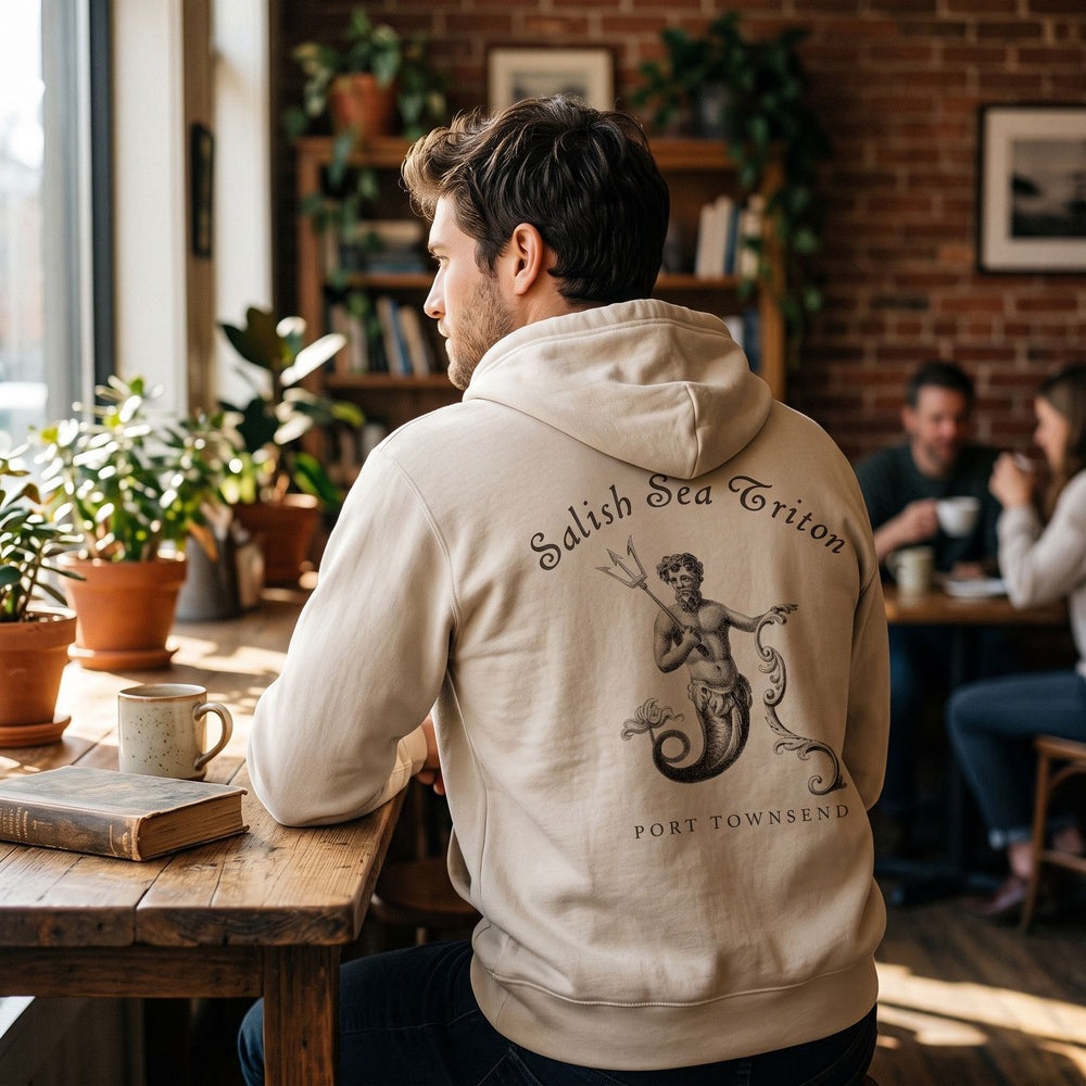 Model wearing a tan heavyweight full-zip hoodie with Triton graphic in a cozy cafe setting.
