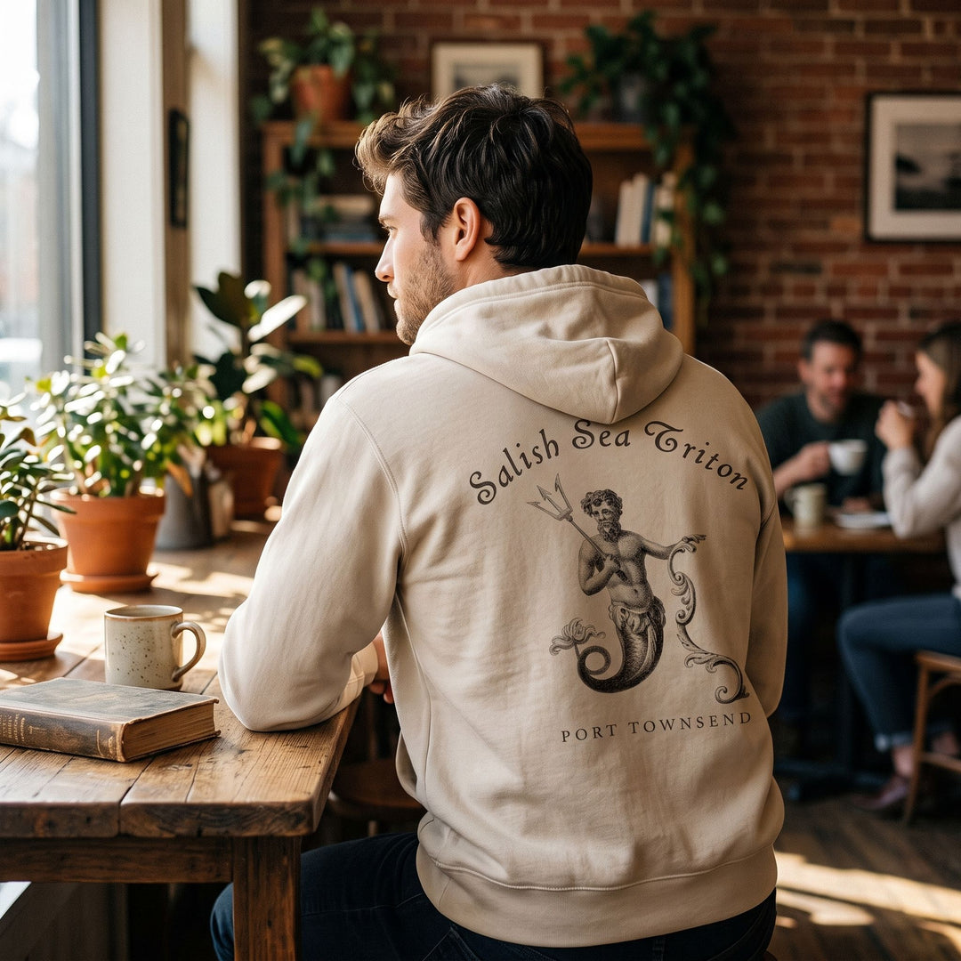 Model wearing a tan heavyweight full-zip hoodie with Triton graphic in a cozy cafe setting.
