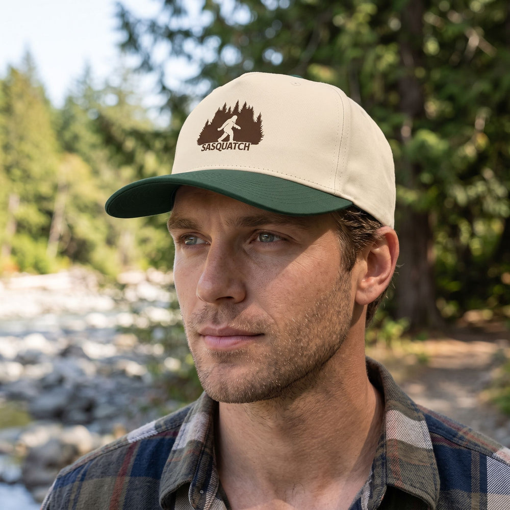 Outdoor scene with model wearing Sasquatch Tracker 5-Panel Cap in natural daylight with greenery, rocks, and water in the background.