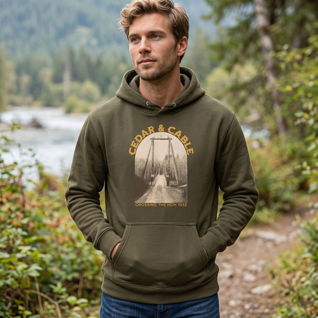 Adult model wearing the Cedar &amp; Cable hoodie outdoors with blurred foliage and water in the background on a sunny day.